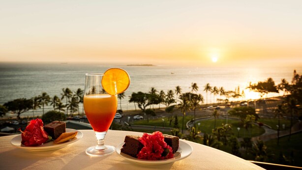 Une table avec un verre de jus d’orange, une boisson rouge et deux assiettes de nourriture, donnant sur une vue côtière avec des palmiers et un coucher de soleil.
