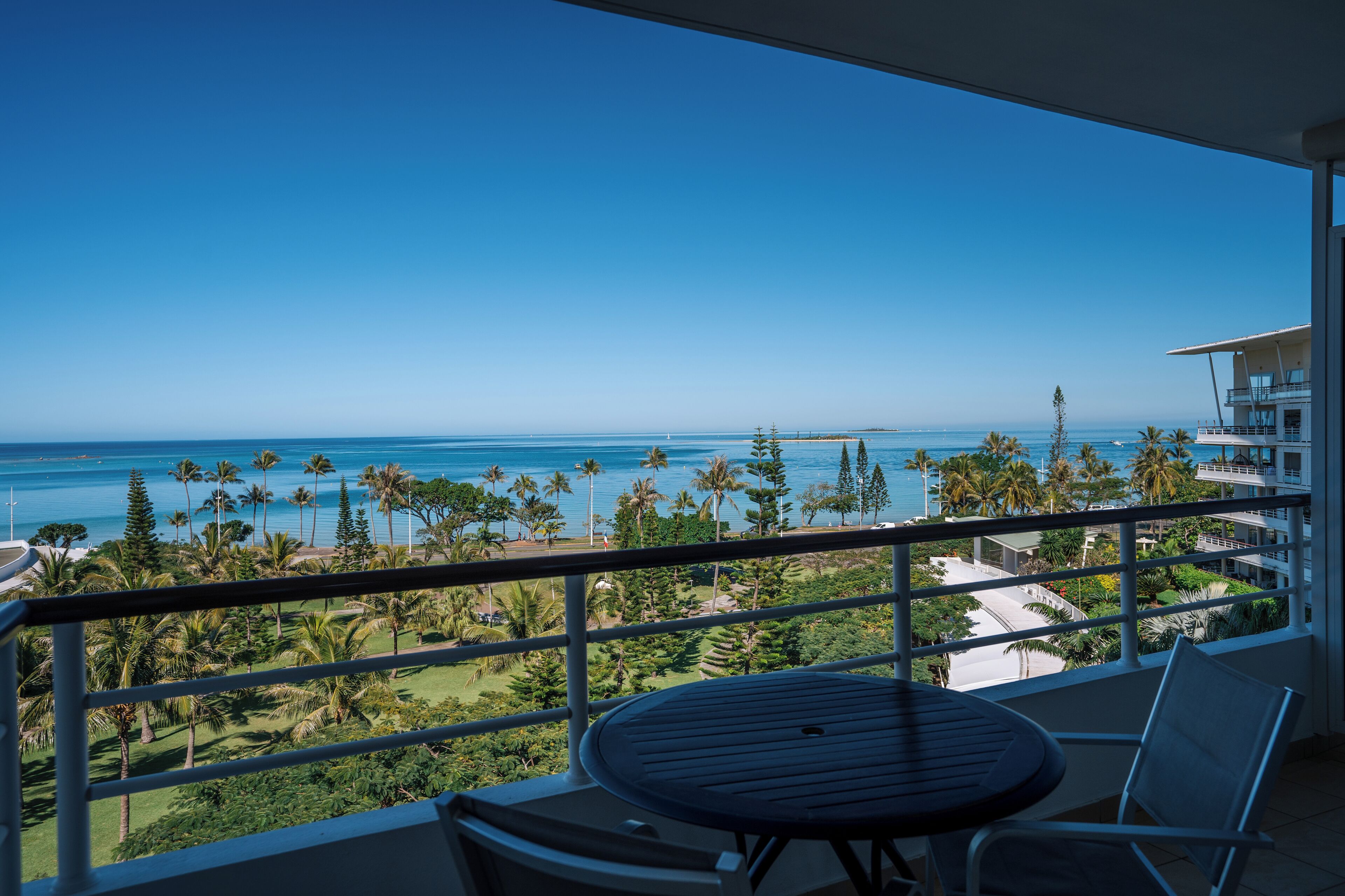 A balcony with a table and chairs overlooking a coastal view with palm trees and a clear sky.