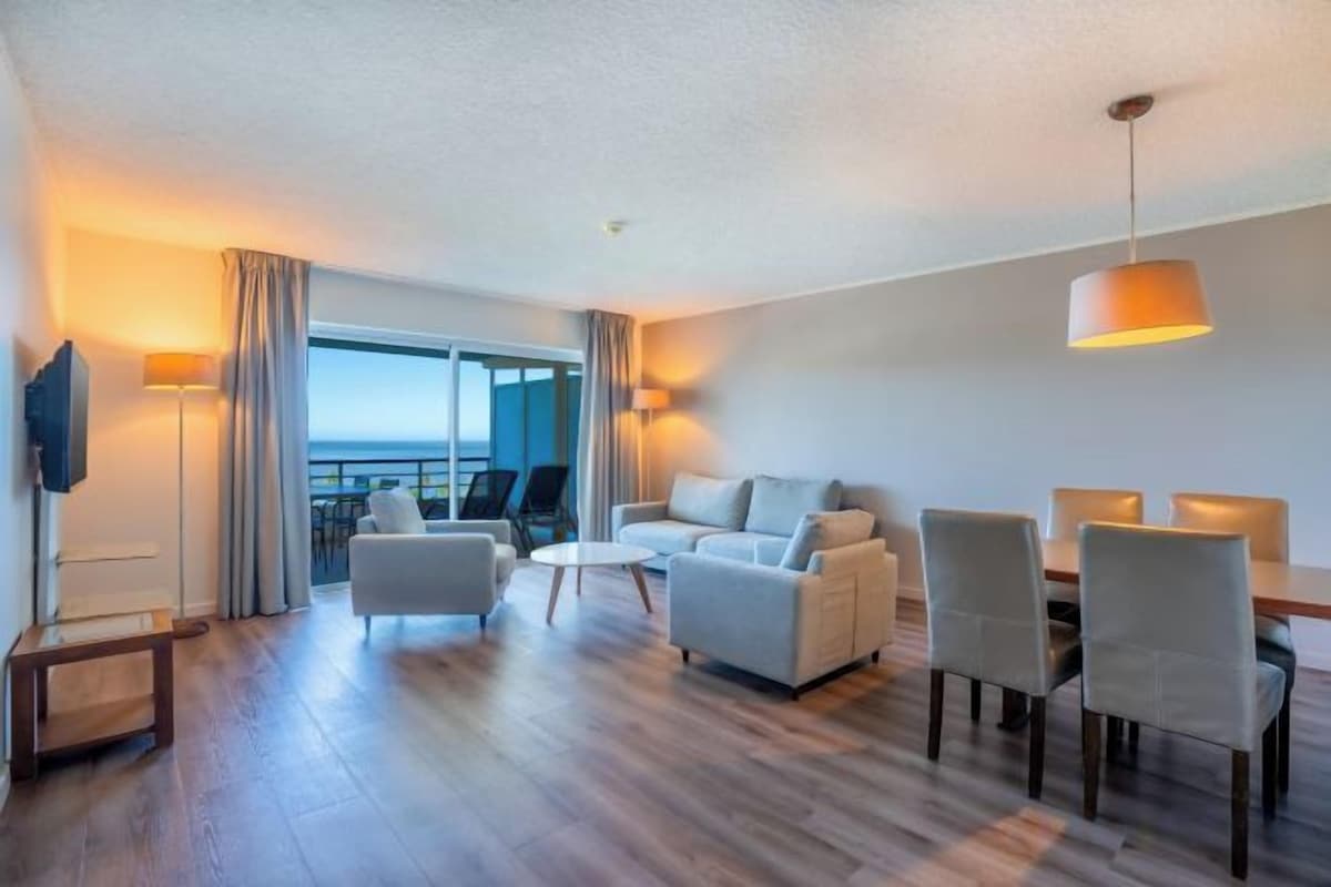 Habitación de hotel con balcón, sofá, mesa de comedor y televisor.