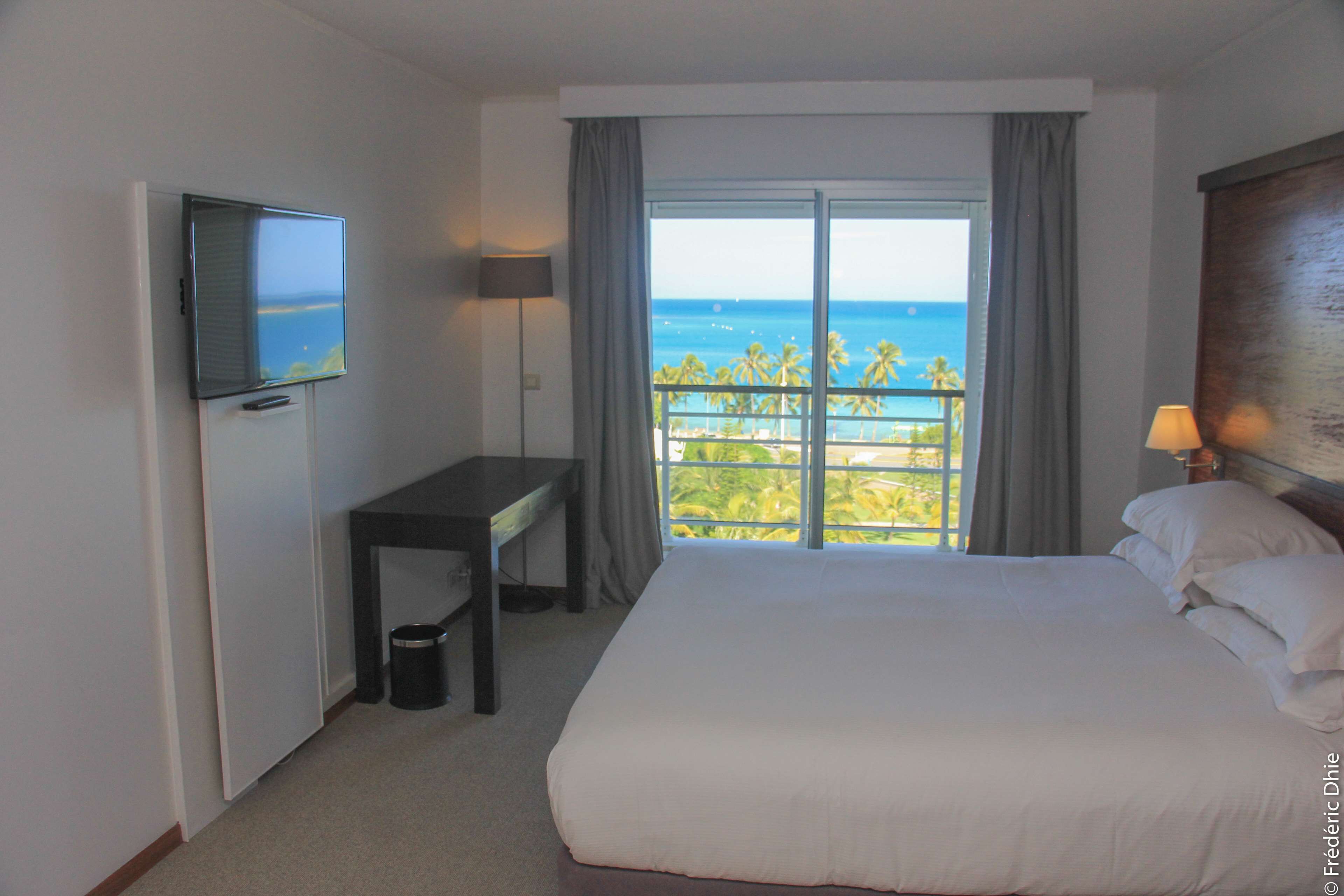 Habitación de hotel con cama, escritorio, televisión y balcón con vista al océano.