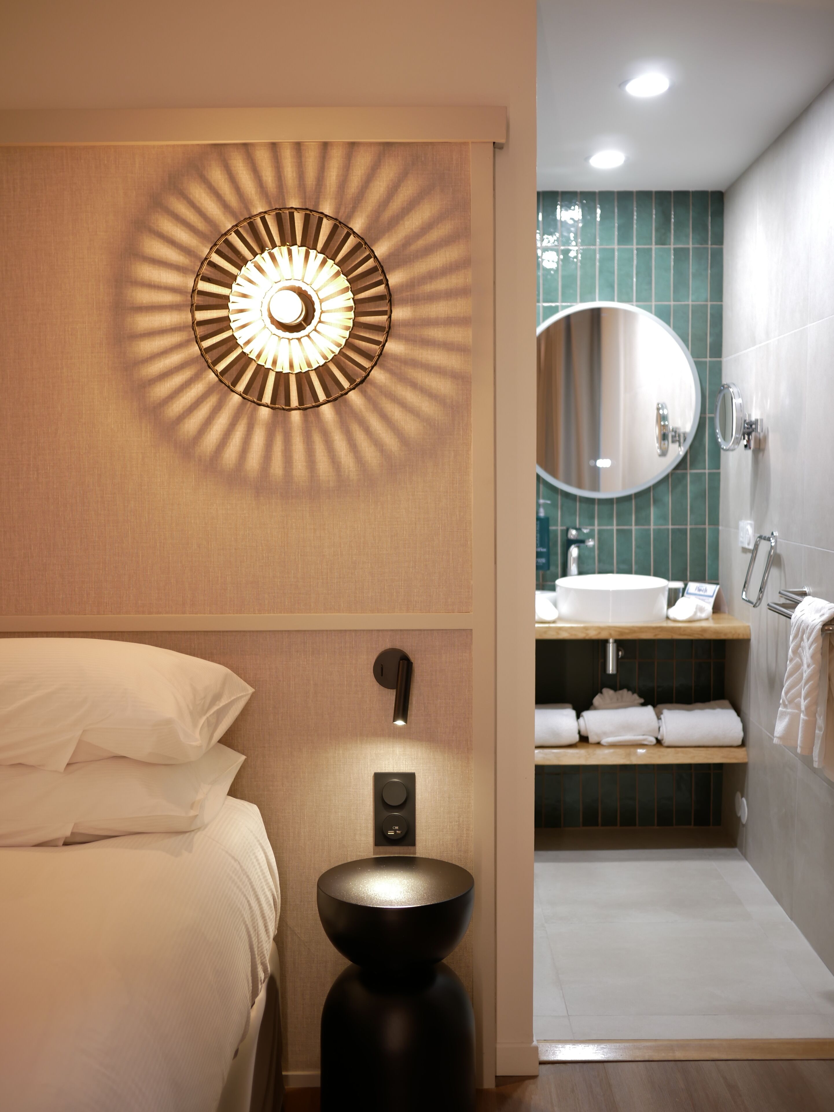 Una habitación de hotel con una cama, una lámpara circular fijada a la pared, un baño moderno con un espejo redondo, un lavabo y toallas ordenadas.