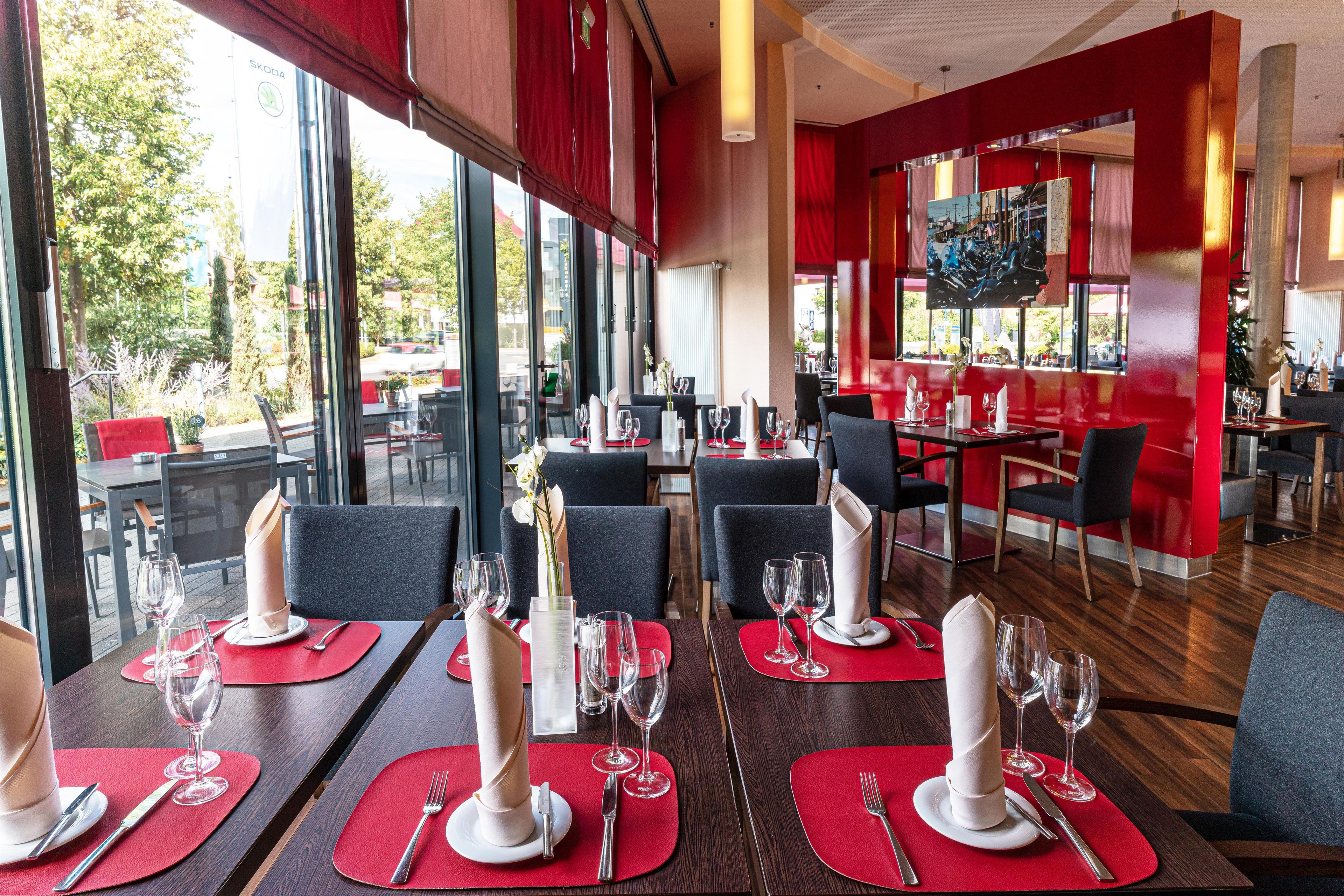 A restaurant with tables set for dining, large windows, and a red accent wall.