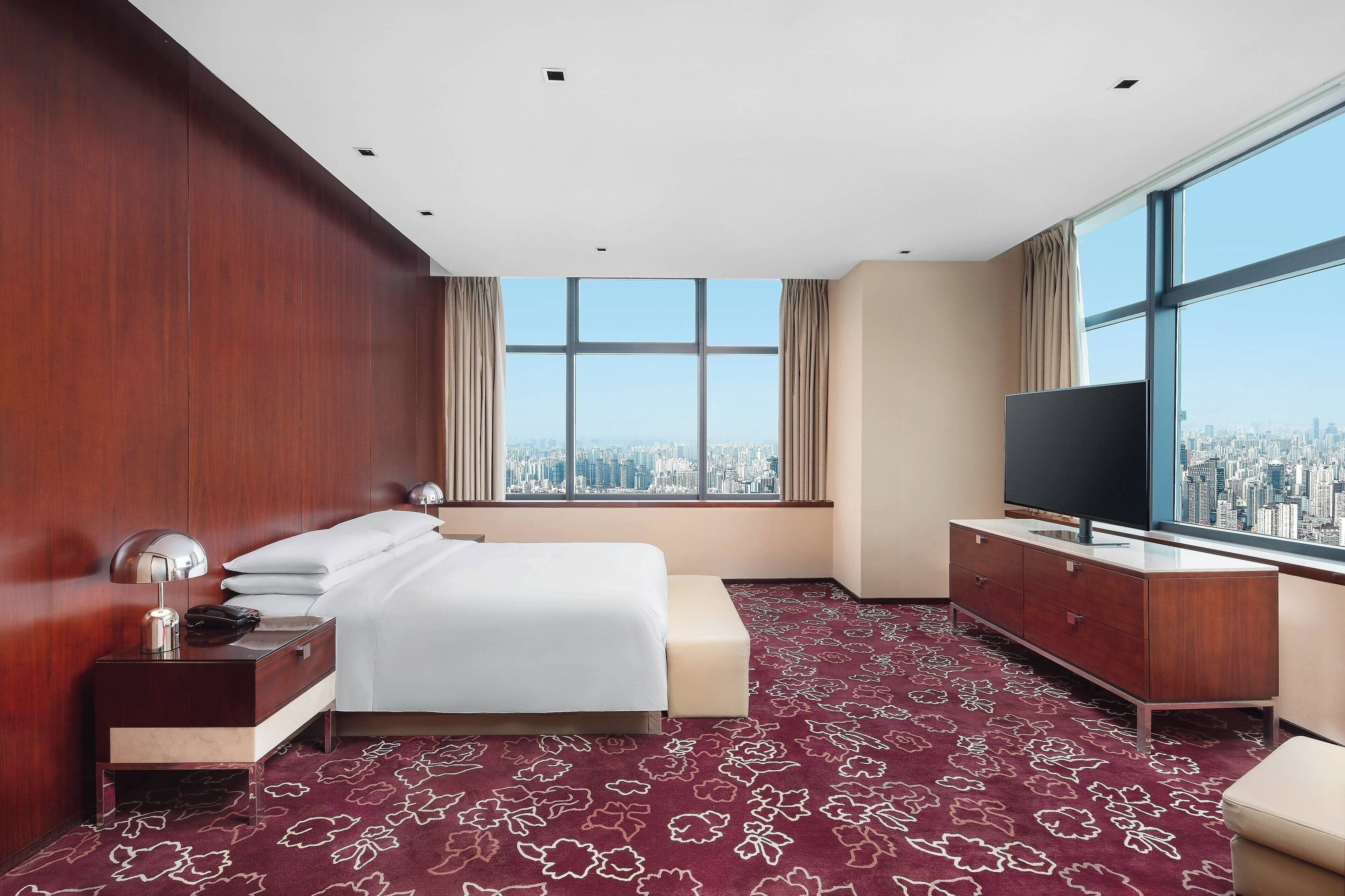 A hotel room with a large bed, a TV, a wooden nightstand, and a view of the city through large windows.