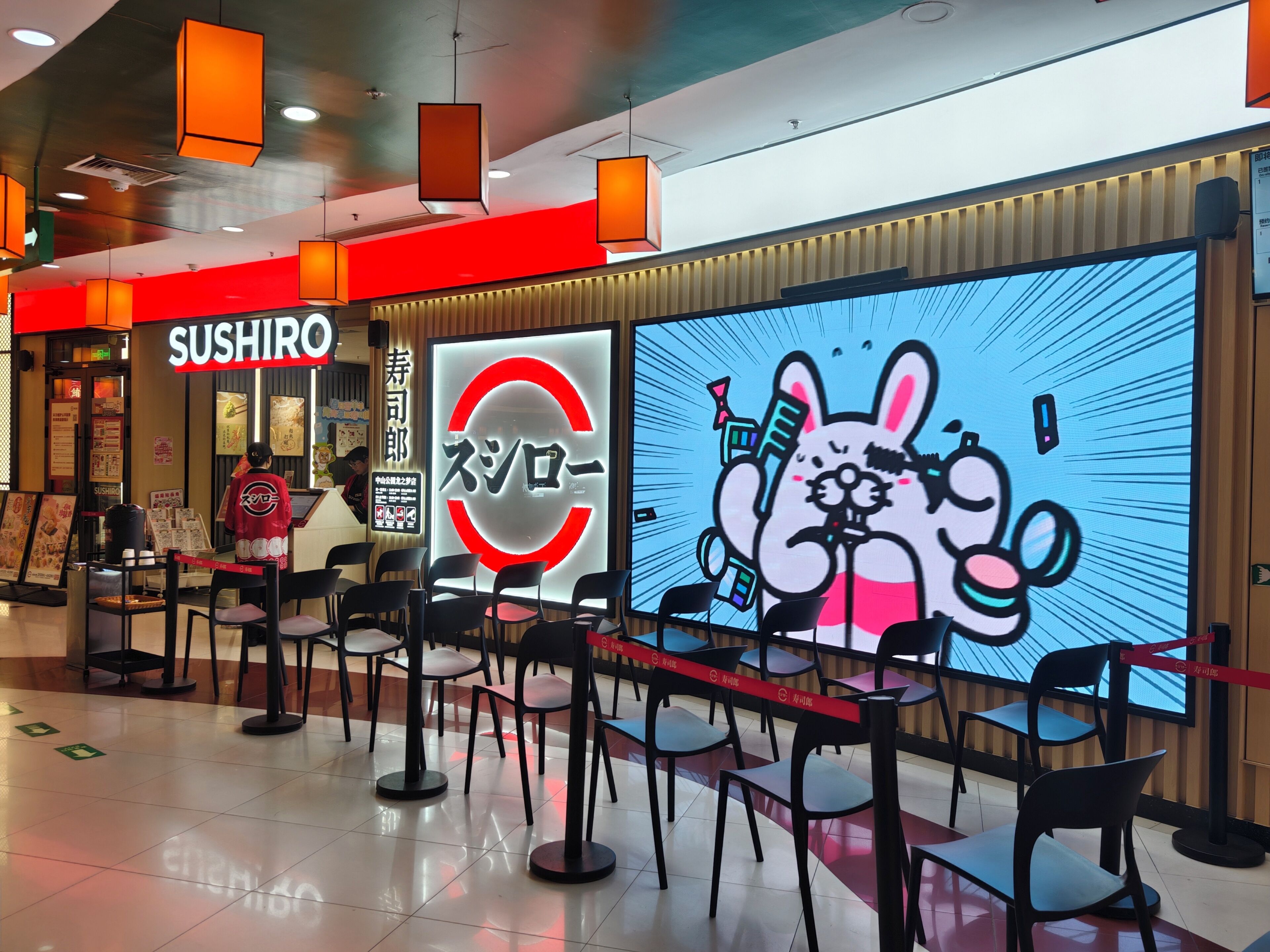 A sushi restaurant with a bright red sign, a large digital screen with a cartoon rabbit, and black chairs arranged in the foreground.