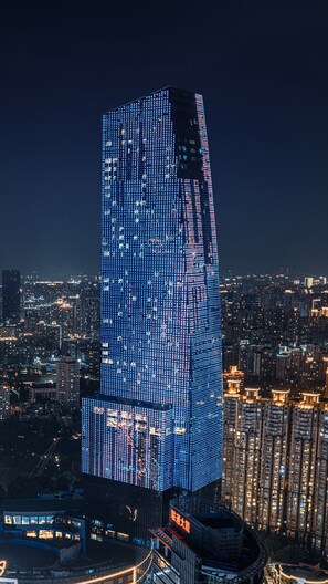 A tall, illuminated skyscraper with a unique, angular design, surrounded by a densely packed urban area at night. - Renaissance Shanghai Zhongshan Park Skyline Hotel (Shanghai)