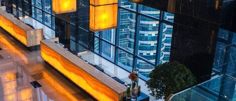 A modern hotel lobby with a glass staircase, a seating area with a vase of flowers, and a view of the cityscape through large windows.