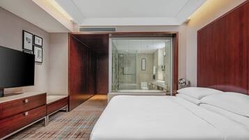 A modern hotel room with a large bed, a TV, a wardrobe, and a glass-enclosed shower area.