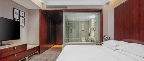 A modern hotel room with a large bed, a TV, a wardrobe, and a glass-enclosed shower area.