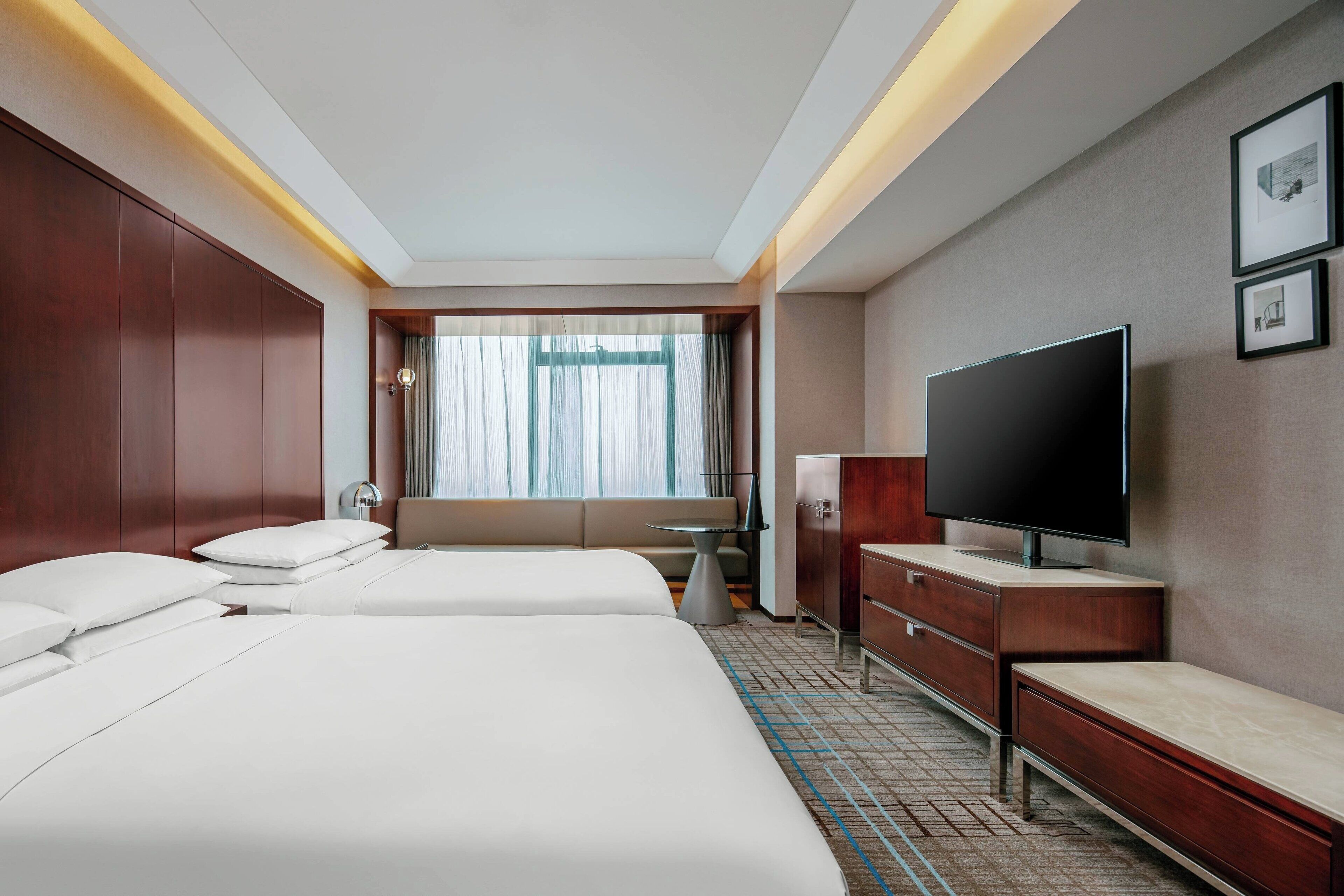 A hotel room with two beds, a TV, a desk, and a window with curtains.