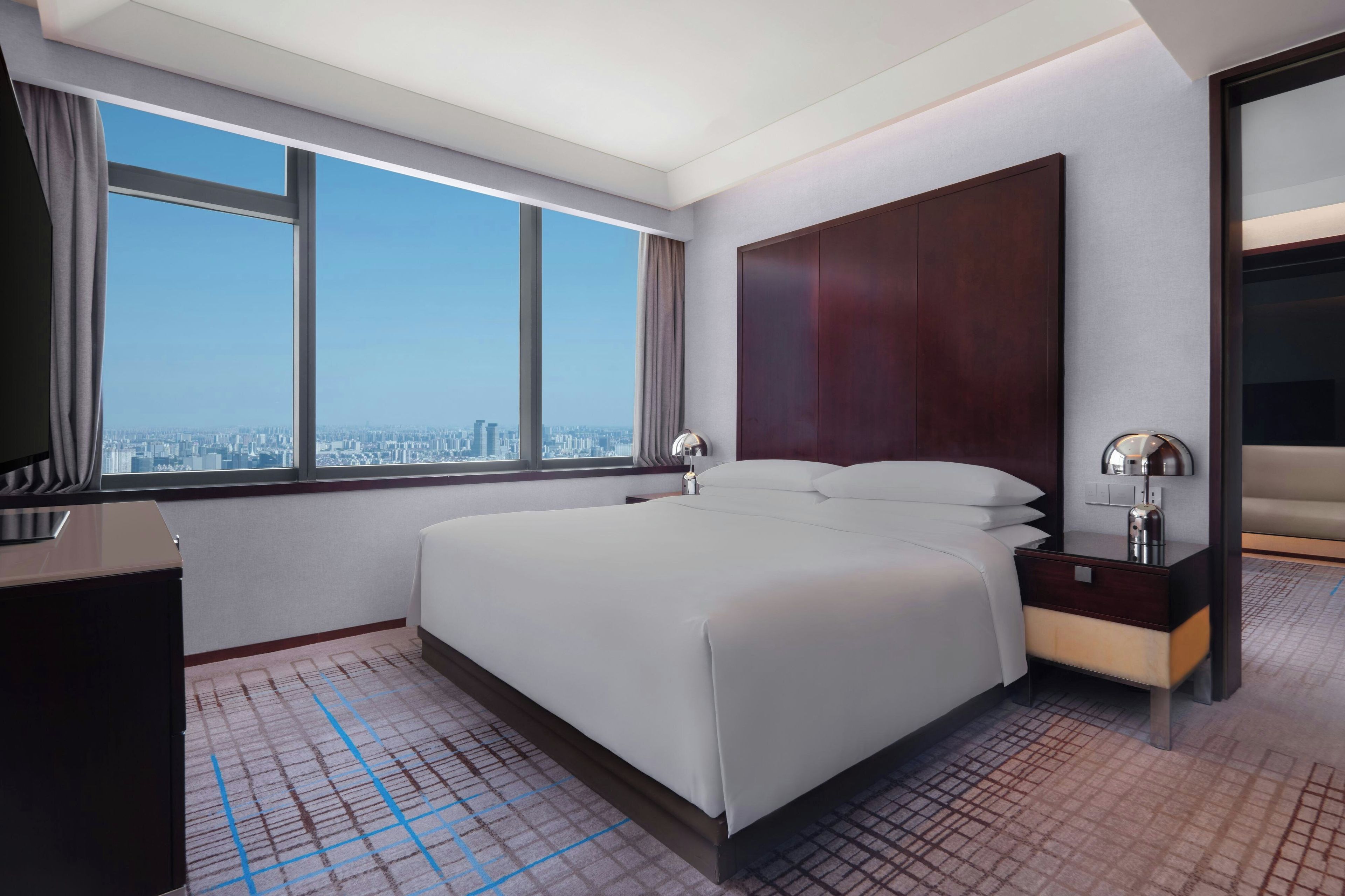 A hotel room with a large bed, a nightstand, a mirror, and a view of the city through a large window.