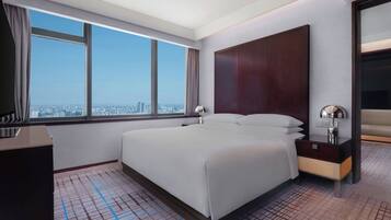 A hotel room with a large bed, a nightstand, a mirror, and a view of the city through a large window.