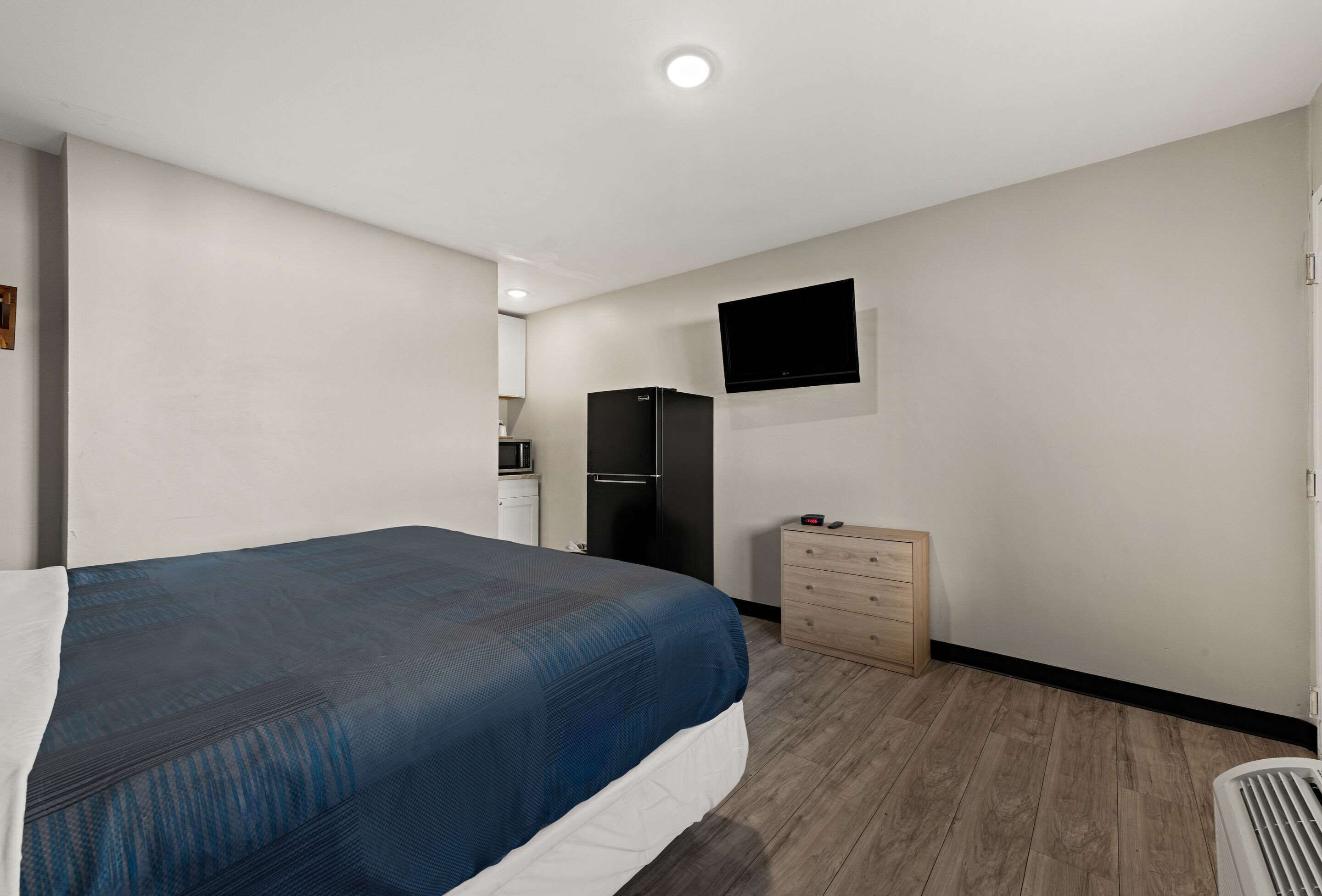 Ein Schlafzimmer mit einem Bett, einer Kommode, einem an der Wand befestigten Fernseher und einem Kühlschrank im Hintergrund.