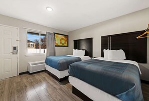 A hotel room with two beds, a large window, a heater, and a painting on the wall.