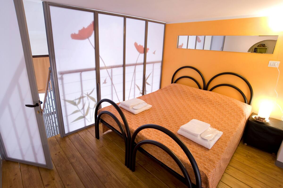 A bedroom with a bed, a nightstand, a mirror, and a sliding door with a view of flowers.