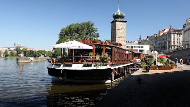 A riverboat docked at a pier with a restaurant on board, a tower in the background, and a cityscape across the water. - Boat Hotel Matylda (Prague)