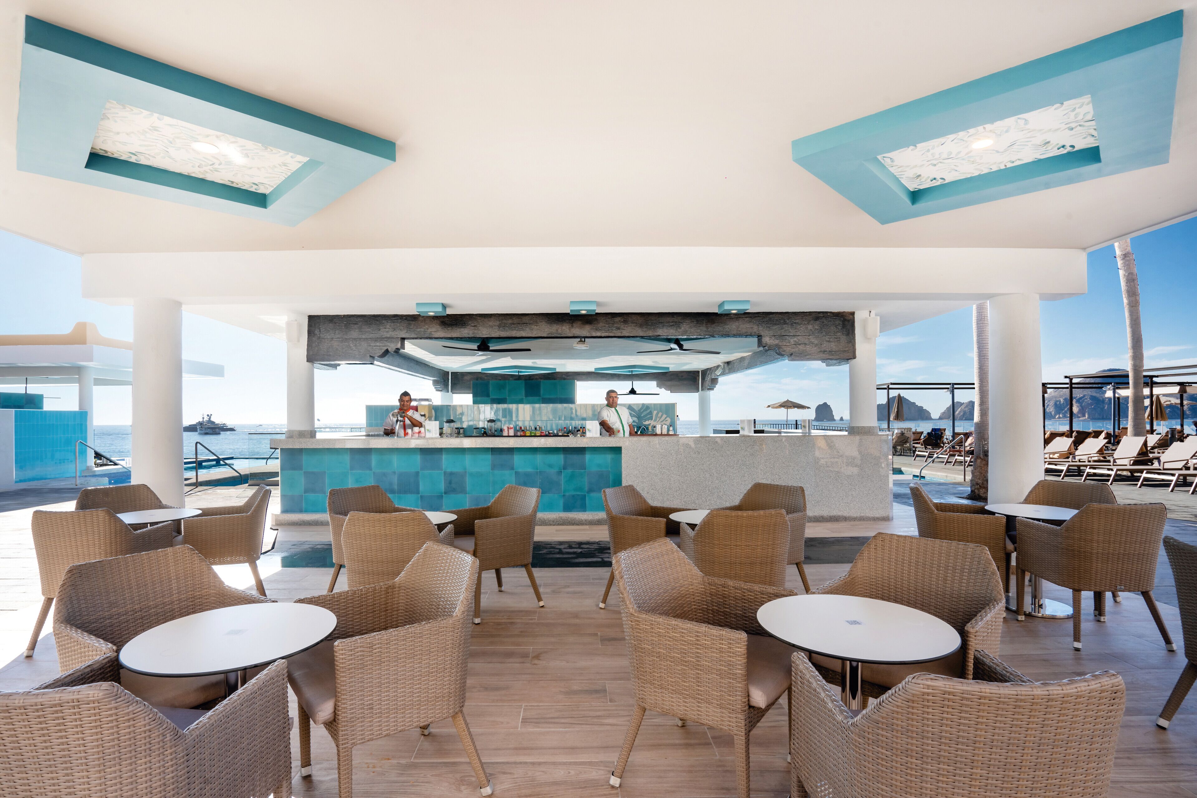 A spacious outdoor lounge area with wicker furniture and a bar in the center, overlooking the sea.