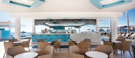 A spacious outdoor lounge area with wicker furniture and a bar in the center, overlooking the sea.