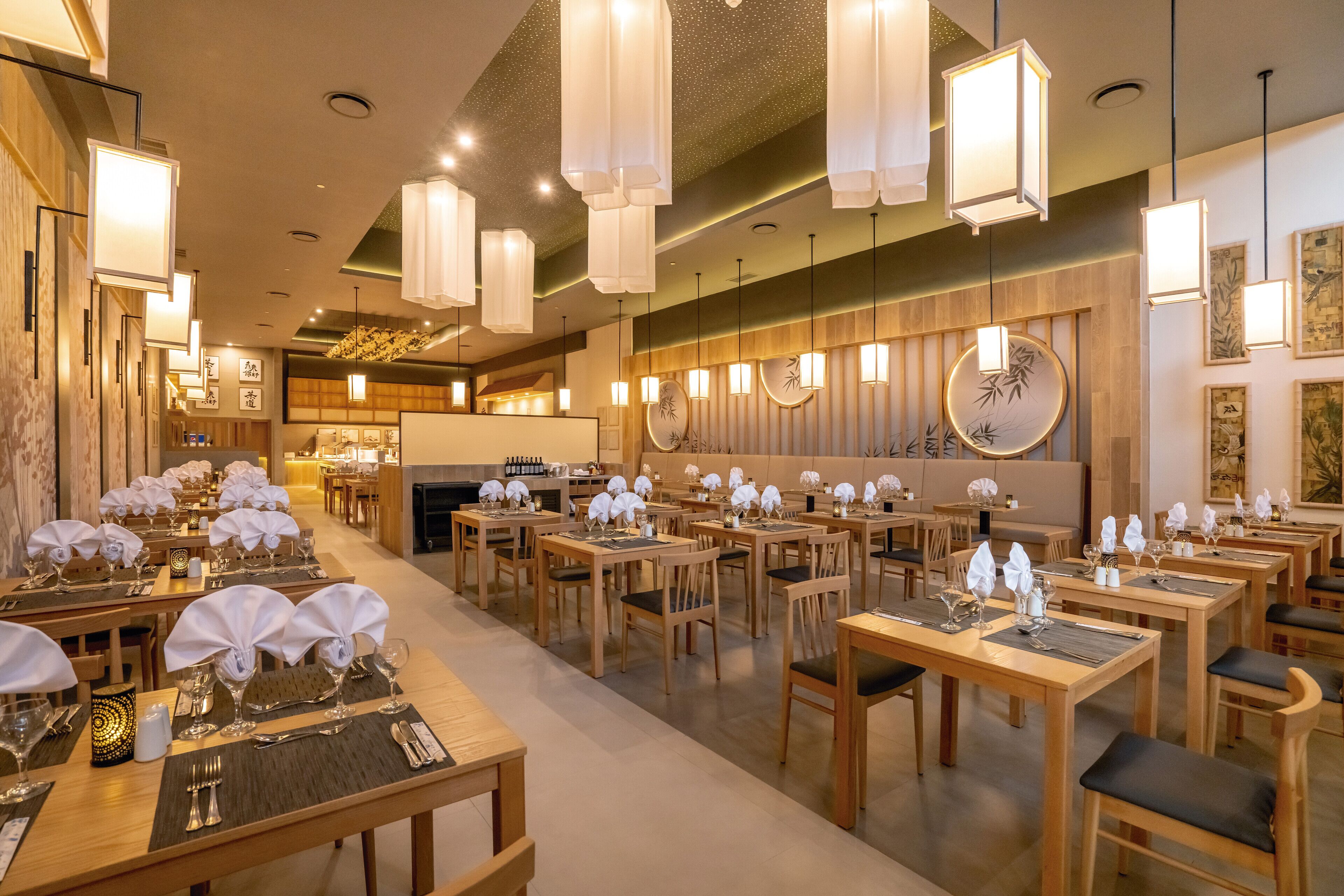 A restaurant with multiple dining tables set with white tablecloths and candles, wooden chairs, and hanging pendant lights.