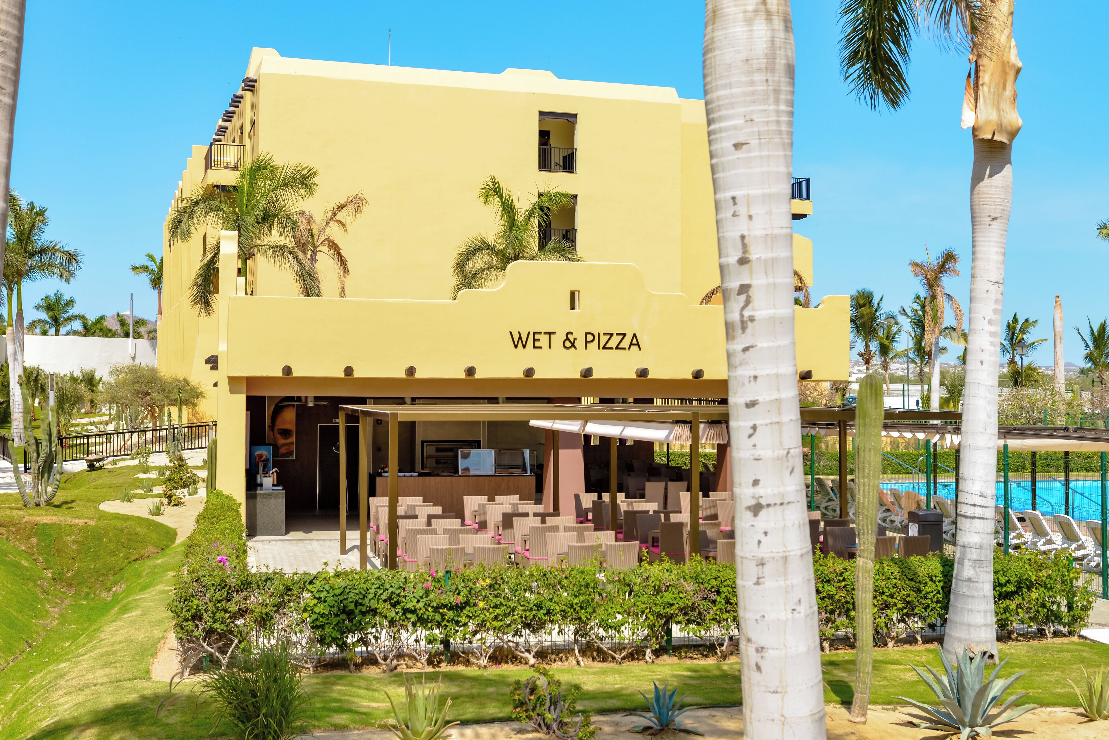 Un bâtiment jaune avec une enseigne indiquant « WET & PIZZA », entouré de palmiers et d’un espace piscine.