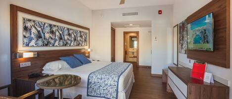 A hotel room with a bed, a desk, a chair, a ceiling fan, and artwork on the walls.
