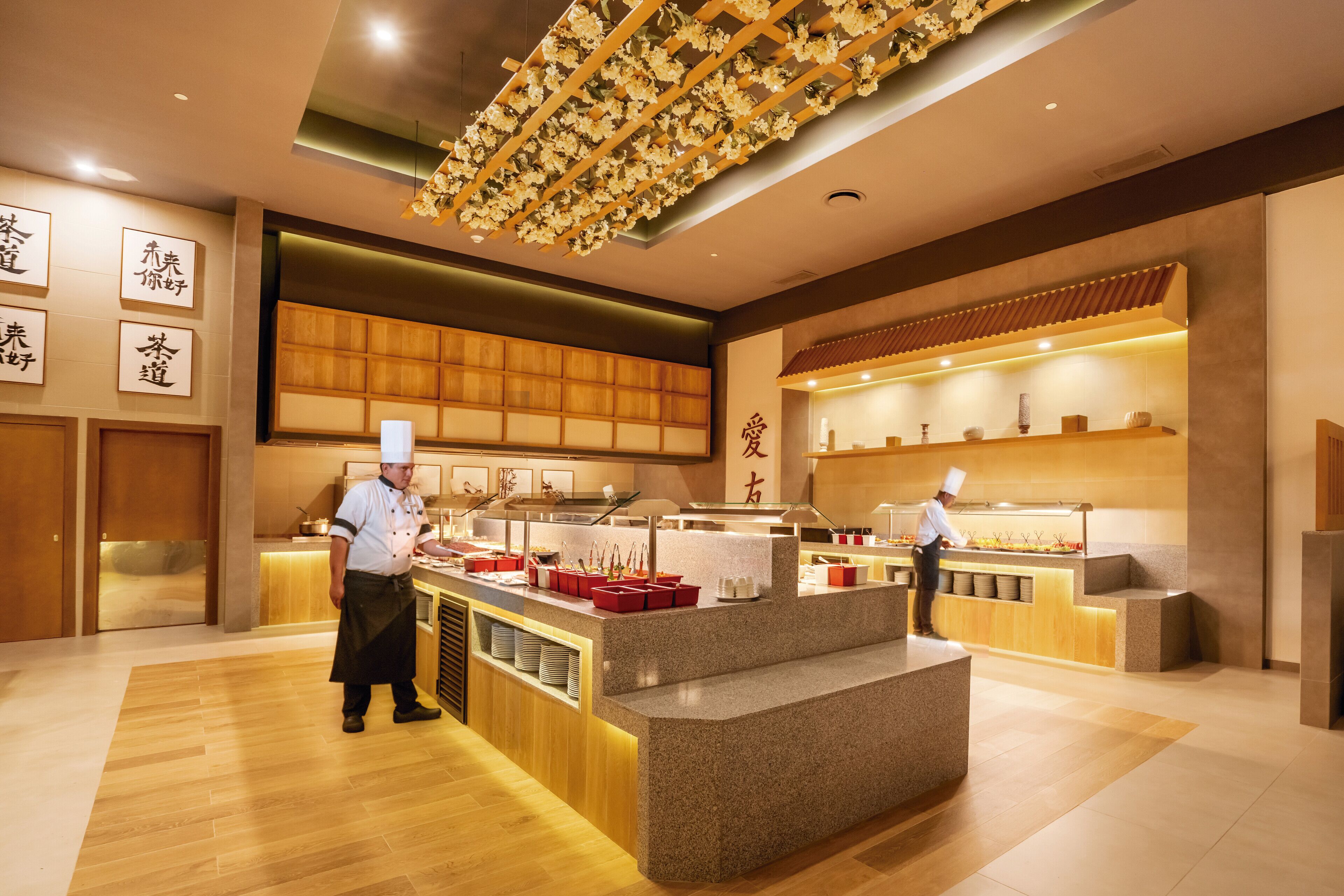 A modern restaurant interior with a central counter, wooden paneling, and a ceiling with floral decorations.