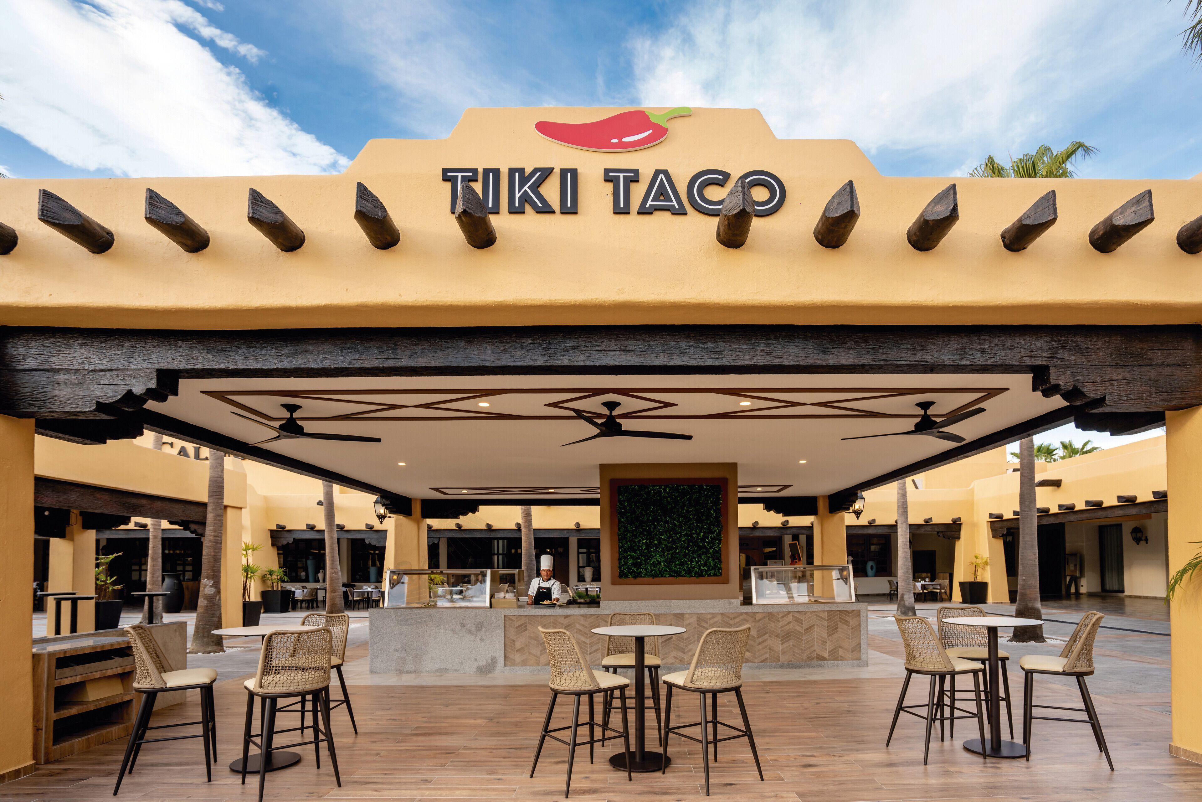 An outdoor dining area with a TIKI TACO sign, wooden tables, wicker chairs, and a ceiling fan.
