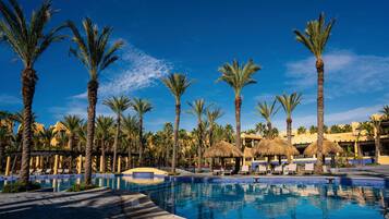 Zona de piscina en un resort con palmeras y sombrillas de paja, rodeada de sillas de descanso y un cielo azul despejado.