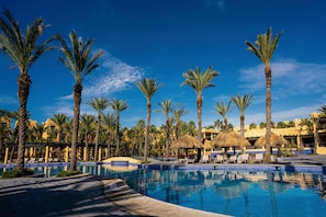 A resort pool area with palm trees and thatched umbrellas, surrounded by lounge chairs and a clear blue sky.