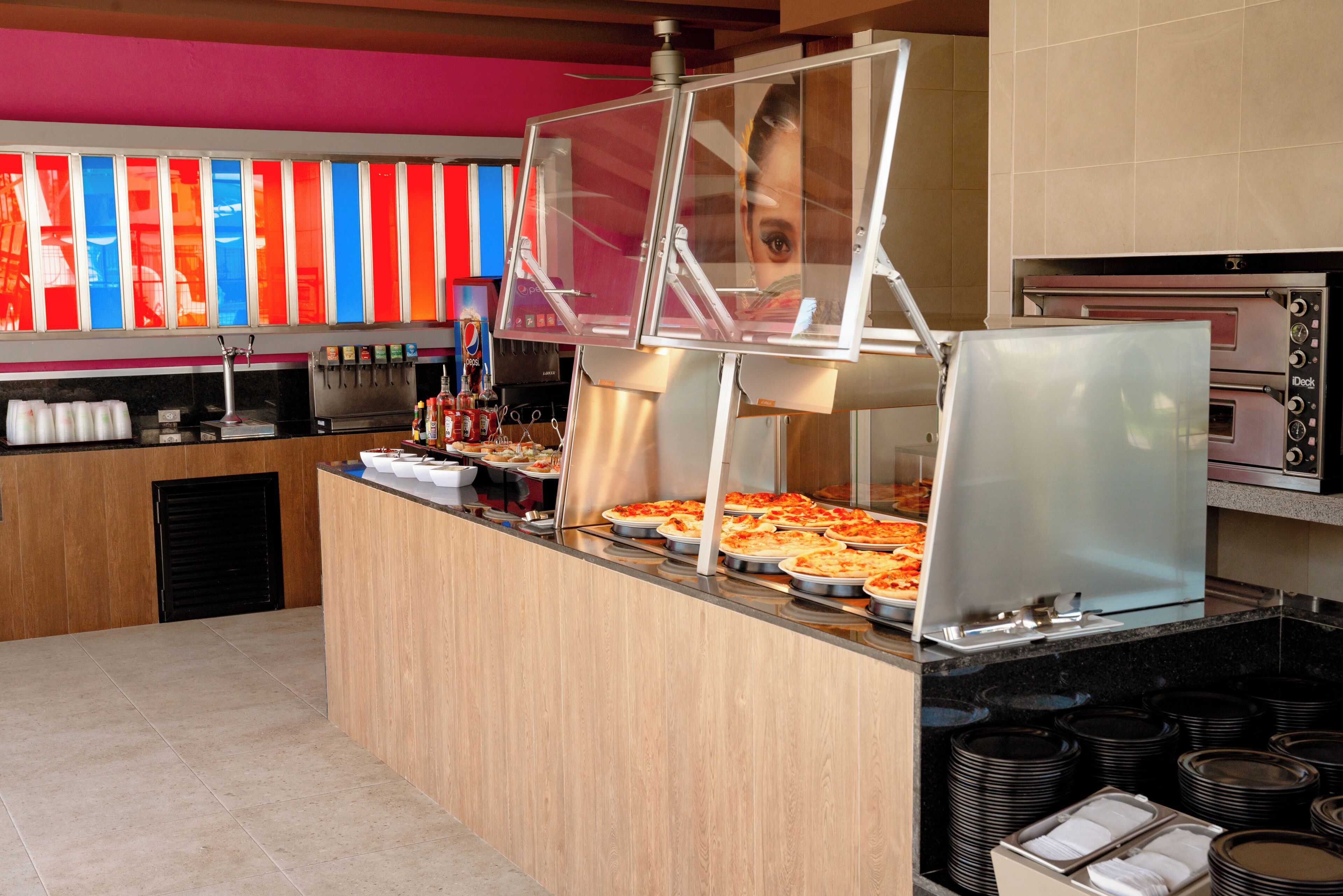 Un espace buffet avec plusieurs plateaux de pizzas, un distributeur de boissons et un four à pizza.
