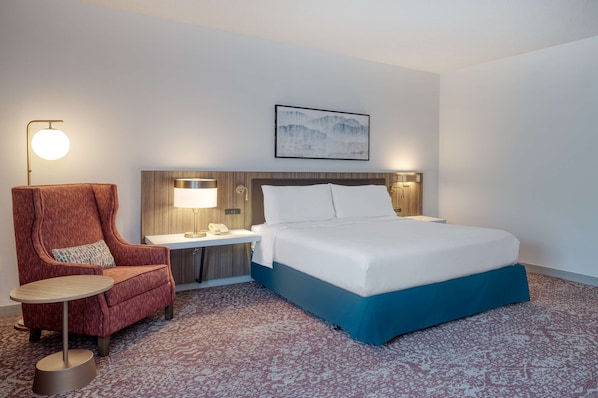 A hotel room with a bed, a red armchair, a small table, a lamp, and a wall-mounted artwork.