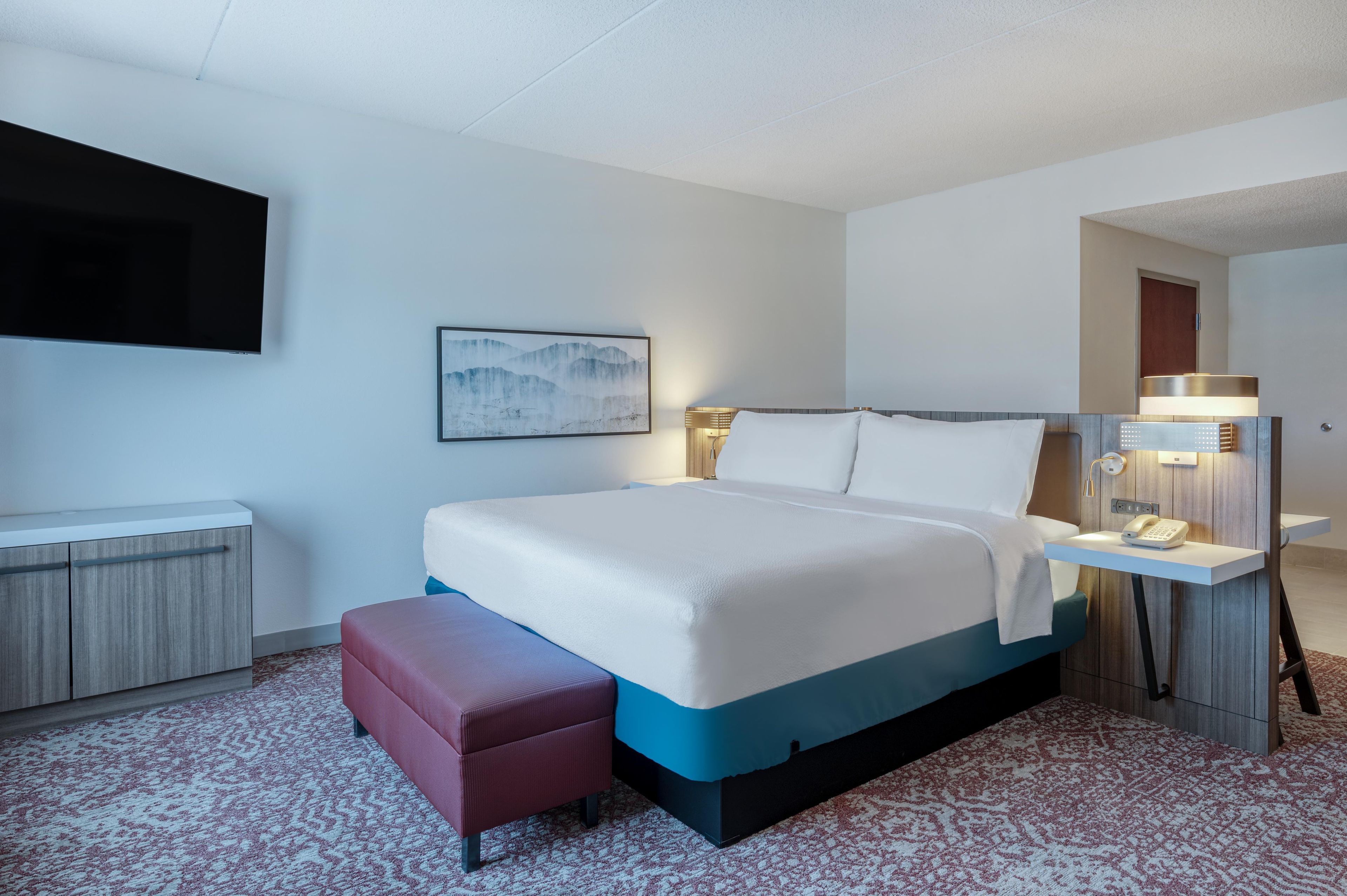 Quarto de hotel com cama, mesa de cabeceira, abajur e televisão fixada na parede.