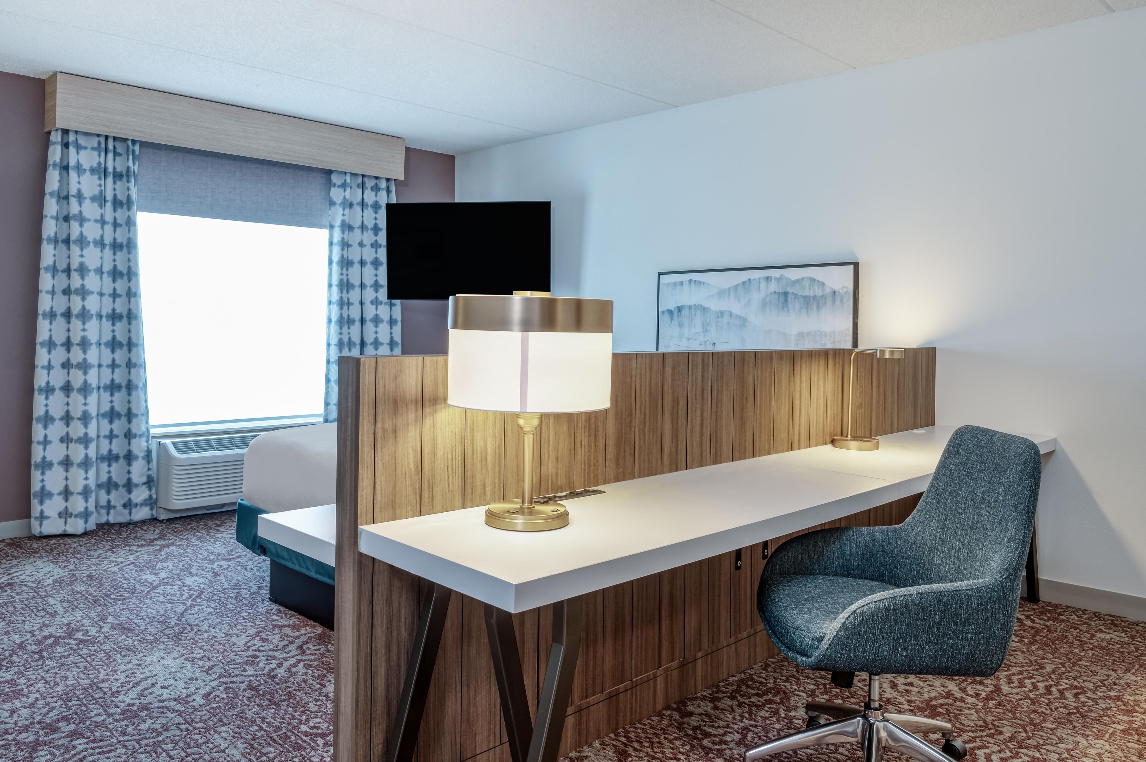 Une chambre d’hôtel équipée d’un bureau, d’une chaise, d’une lampe, d’une télévision et d’une œuvre d’art au mur.