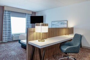 A hotel room with a desk, chair, lamp, TV, and artwork on the wall. - Hilton Garden Inn Clarksburg Bridgeport (Clarksburg)