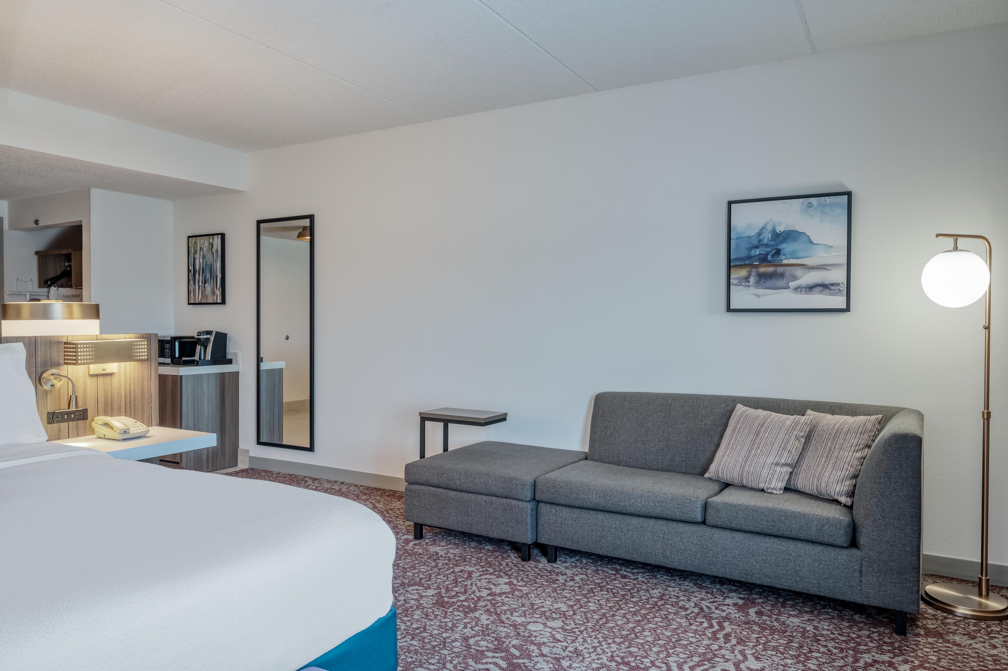 Habitación de hotel con cama, sofá gris, una mesita, lámpara, cuadro y espejo.