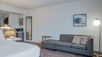 Habitación de hotel con cama, sofá gris, una mesita, lámpara, cuadro y espejo.