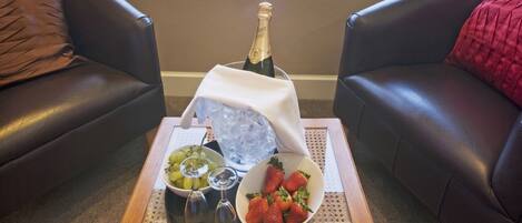A room with a view of a lake and mountains, a table with a bottle of champagne, a bowl of strawberries, and a bowl of grapes.