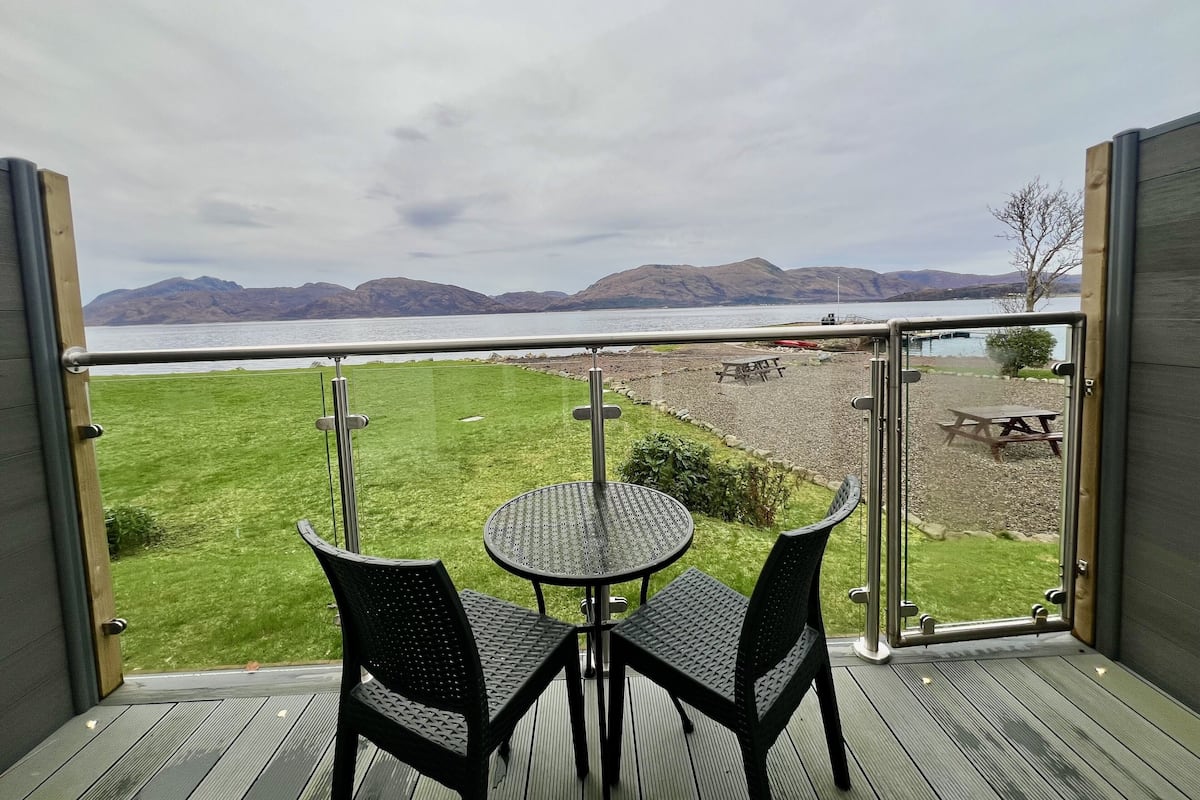 Un balcon avec une table et des chaises, donnant sur un espace verdoyant où se trouve une table de pique-nique, et avec des montagnes visibles au loin.
