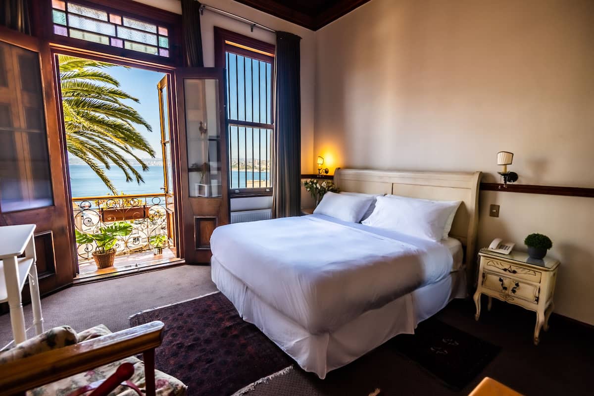 A hotel room with a large bed, a nightstand, a chair, and a view of the sea through an open door.