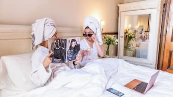 Two people in white robes reading a magazine on a bed with a laptop and a smartphone.