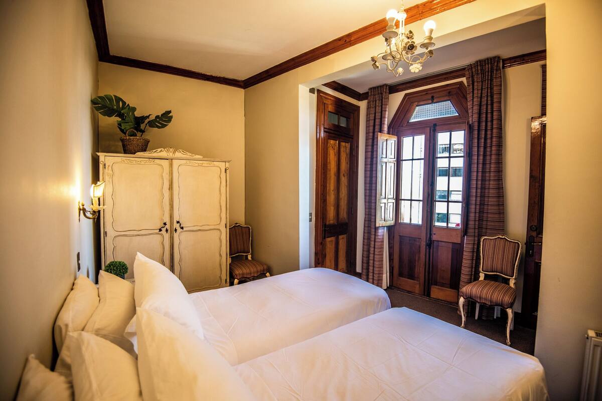 A hotel room with two beds, a wooden wardrobe, a chandelier, and a view of the outside through a window with curtains.
