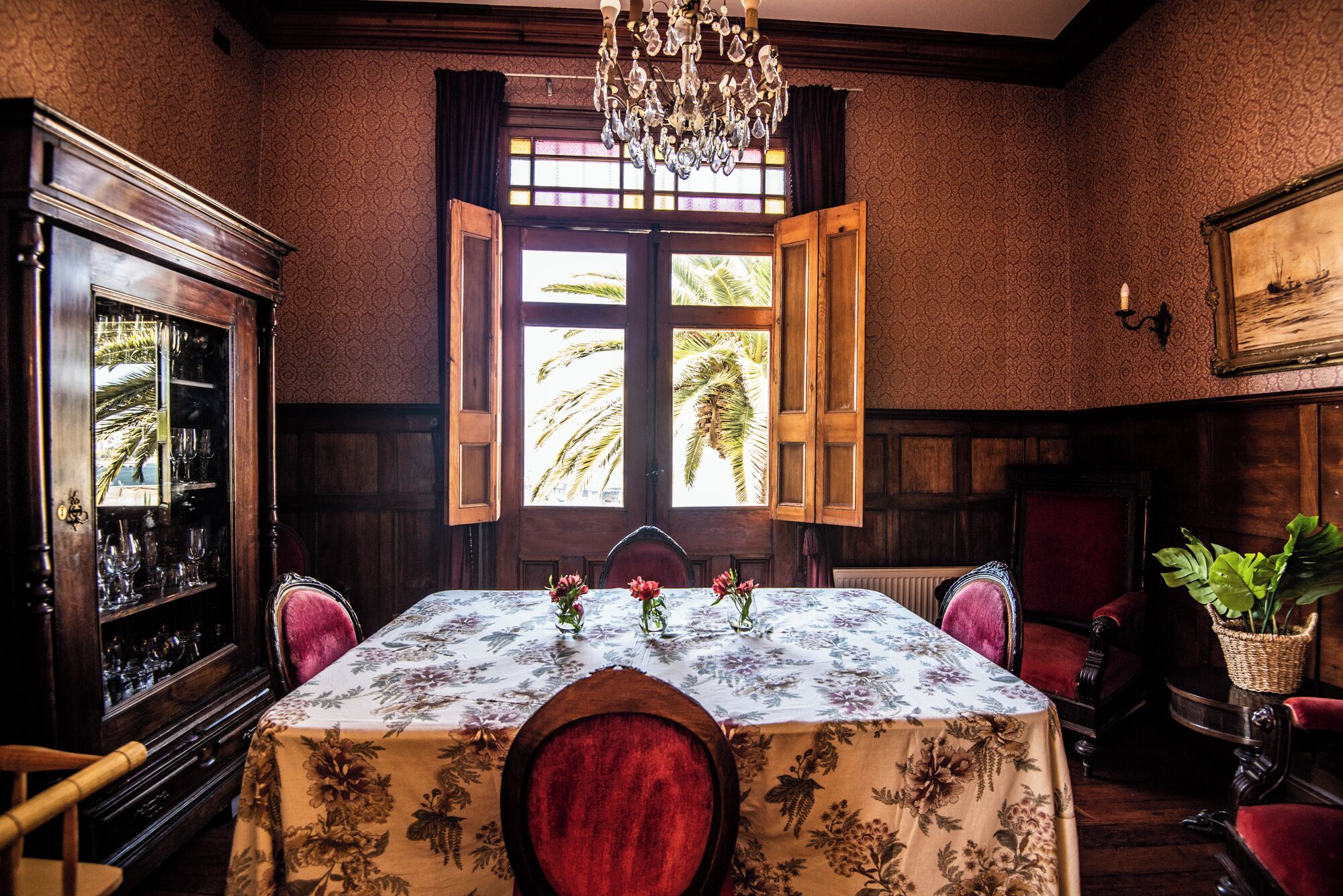 Comedor con una mesa lista para cuatro personas, un candelabro, un ventanal con contraventanas abiertas y un aparador con cristalería.
