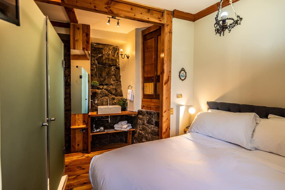 A bedroom with a bed, wooden ceiling beams, a stone wall, a sink, and a mirror.