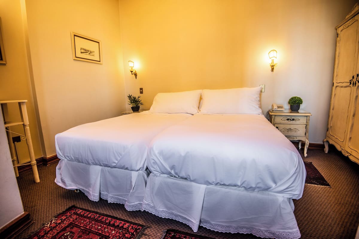 A double bed with white linens, a bedside table with a lamp, a small potted plant, and a framed picture on the wall.