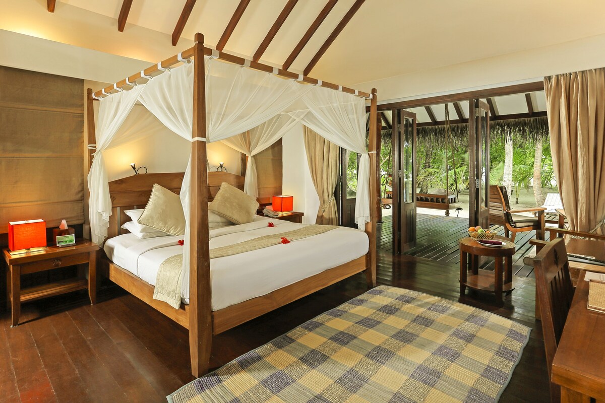 Un lit à baldaquin avec des colonnes aux quatre coins, une tête de lit en bois, des tables de chevet avec des lampes, un tapis à carreaux et une vue sur un jardin tropical par de grandes portes vitrées.
