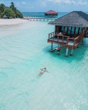 Ein tropischer Strand mit klarem, türkisblauem Wasser, eine Bungalowanlage auf Stelzen und eine Person beim Schwimmen.
