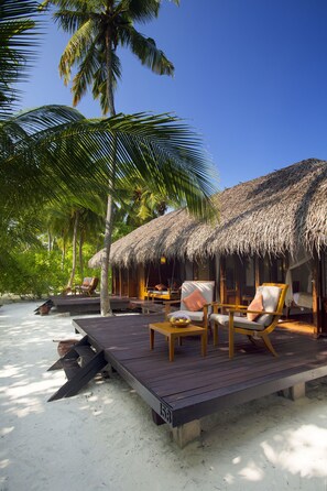 Un resort frente a la playa con cabañas de techo de paja, asientos en terraza de madera y palmeras.