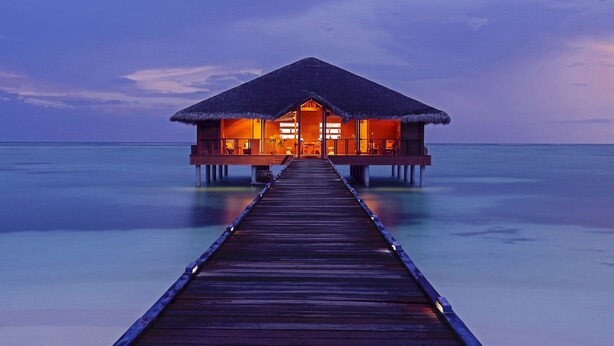 A wooden overwater bungalow with a thatched roof, connected to the mainland by a wooden pier.
