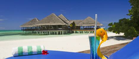 A beachside lounge with a tropical drink, a striped towel, and overwater bungalows in the background.