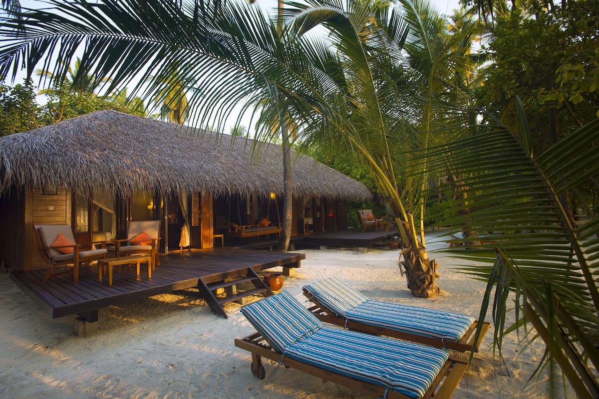 Una cabaña frente a la playa con techo de paja, terraza de madera y sillones de descanso.