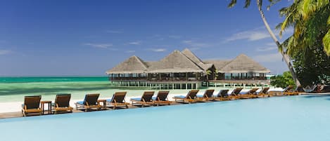 A poolside view with lounge chairs, a clear blue sky, and a resort building with thatched roofs.